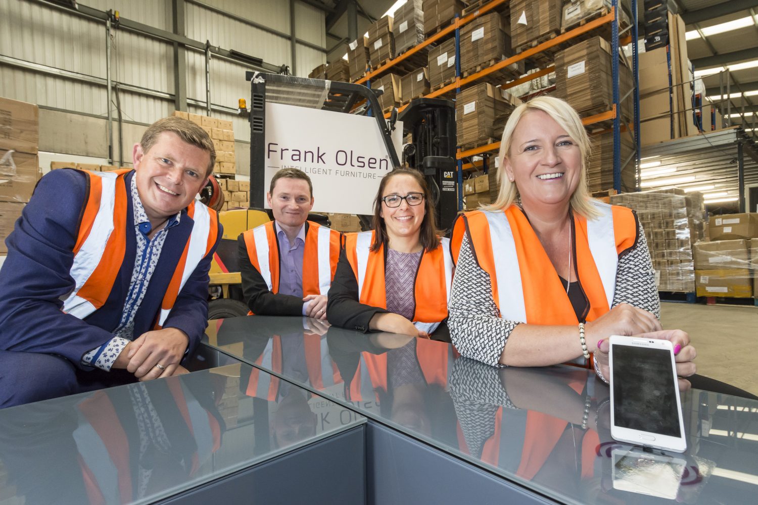 Four women standing in a factory with Frank Olsen signage behind them. They are all wearing high vis jackets. There are two women in the group and the woman to the far right has blonde hair and standing next to her is a woman with dark hair wearing glasses.