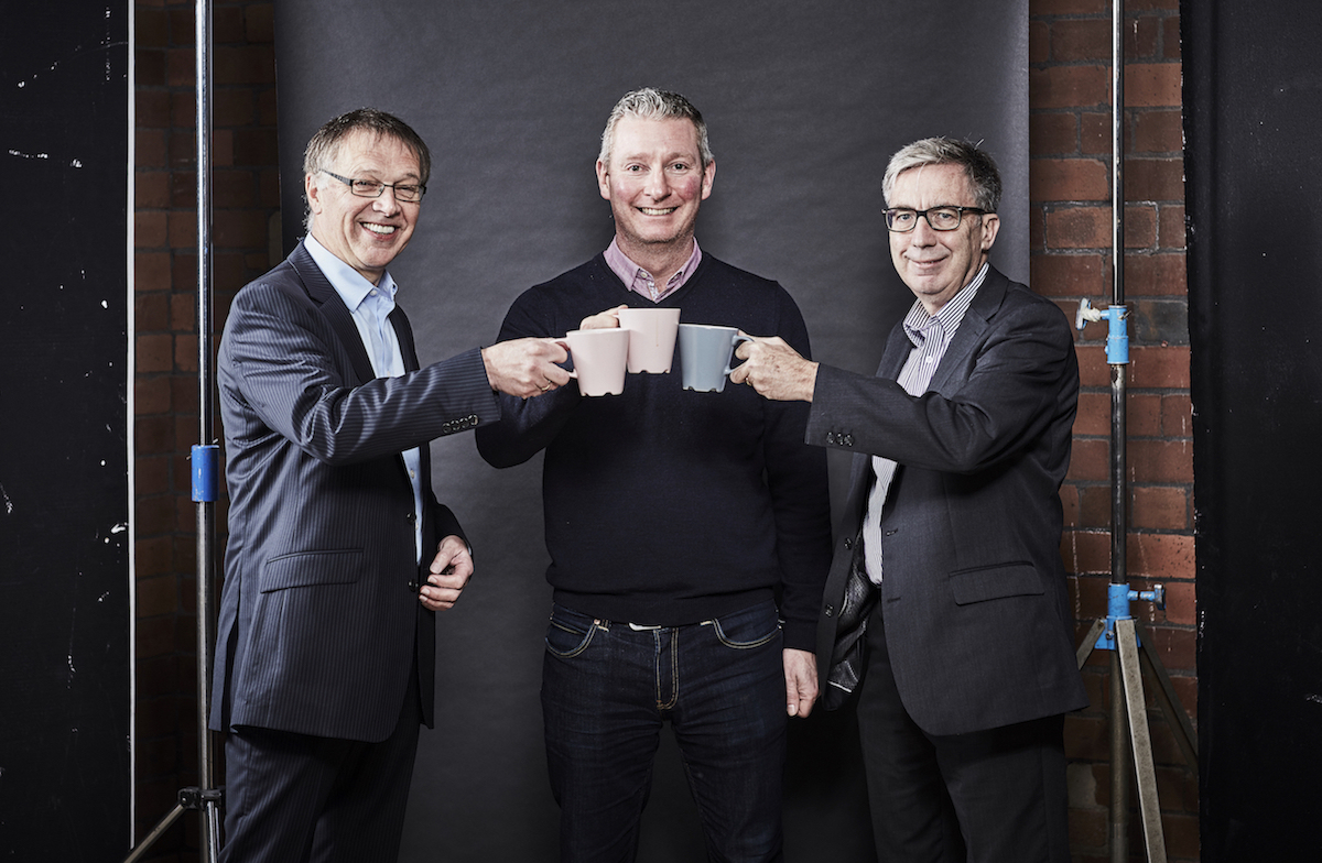 Three men in an office all handing mugs standing behind a brick wall.