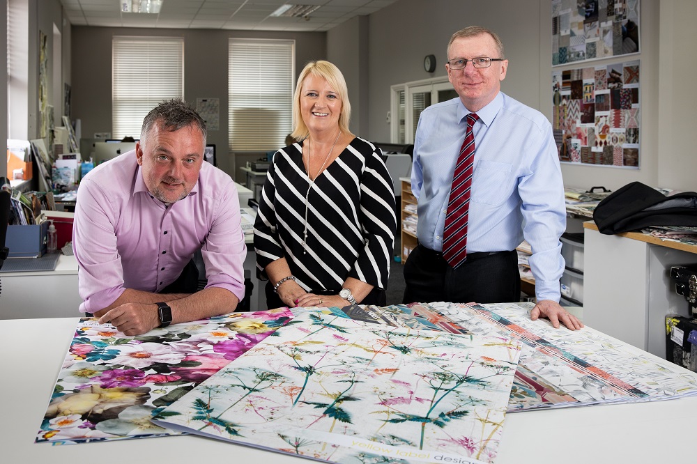 Three people in a design studio standing behind a white desk. There is a woman in the middle of the group with blonde hair wearing a black and white striped jumper. The man to the right is wearing a light blue shirt, has a red stripey tie and has grey hair and glasses. The remaining man is wearing a pink shirt and has his hands on the table.