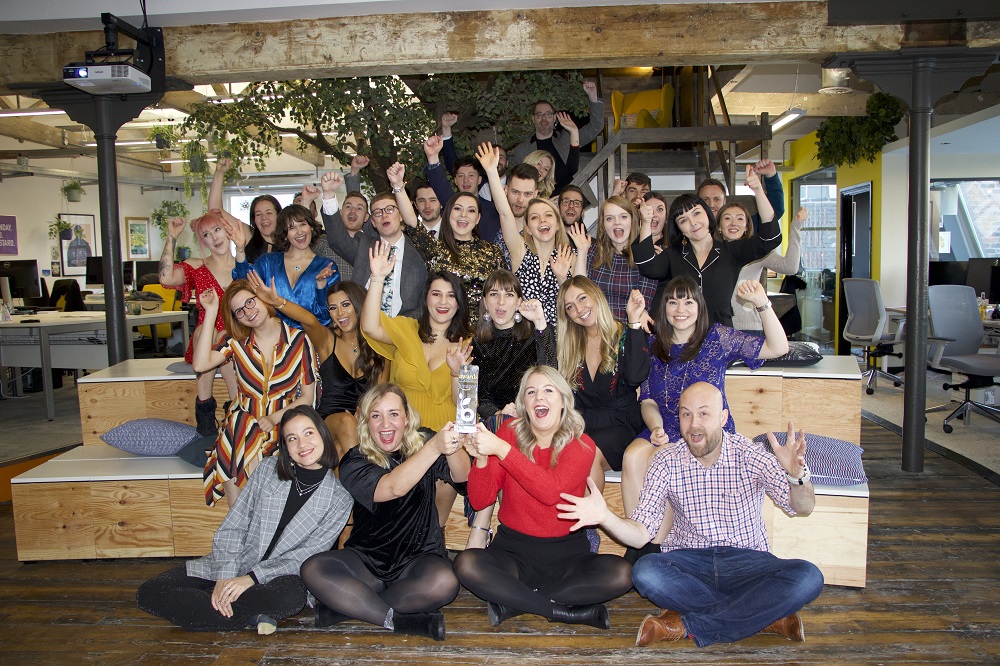 Large group of people in a trendy office. A few people have their arms up in the air and are looking excited.
