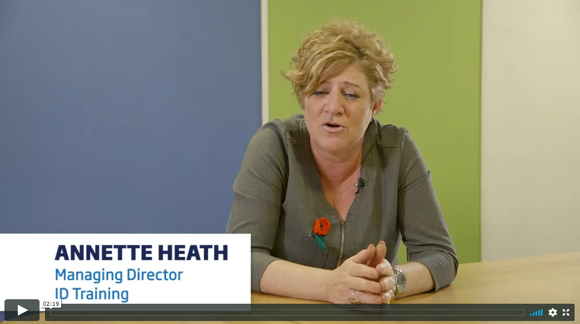 One lady named Annette Heath is sitting behind a desk with short blonde curly hair. She is wearing a khaki dress and there is a remembrance poppy attached to the dress. Behind her the wall is painted in three sections, blue, lime green and pale green. She is Managing Director of JD Training.