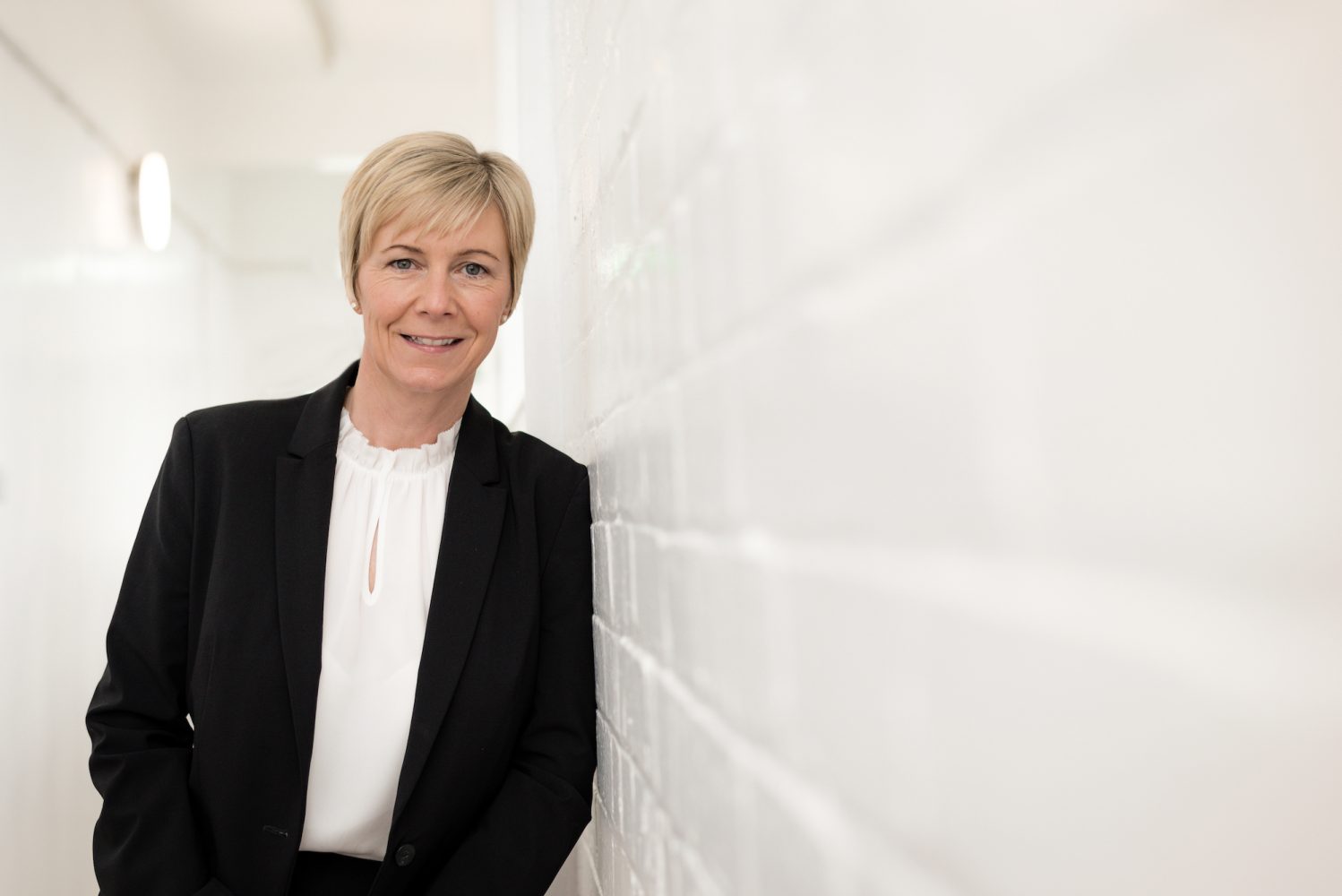 Lady with short blonde hair wearing a black jacket and a white shirt leaning against a white brick internal wall.