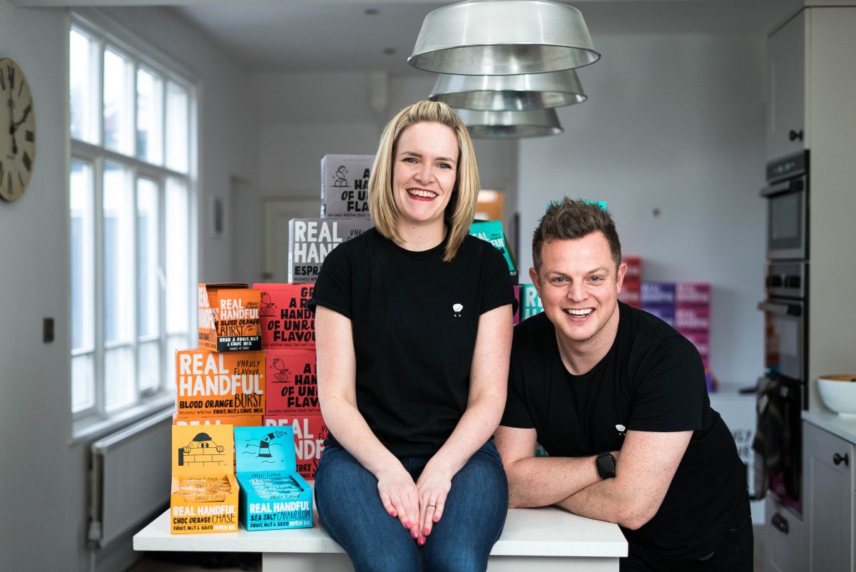 Blond haired lady on a table surrounded by her point of sale and products. She is wearing a black tee shirt and jeans and to the right of her is a young man wearing a black tee shirt and black trousers.