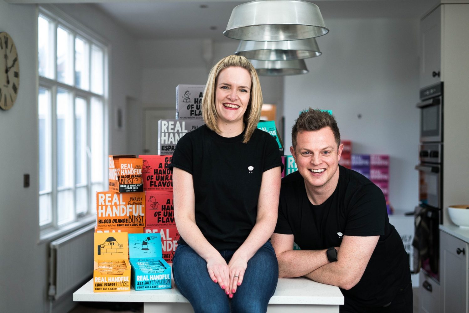 Blond haired lady on a table surrounded by her point of sale and products. She is wearing a black tee shirt and jeans and to the right of her is a young man wearing a black tee shirt and black trousers.