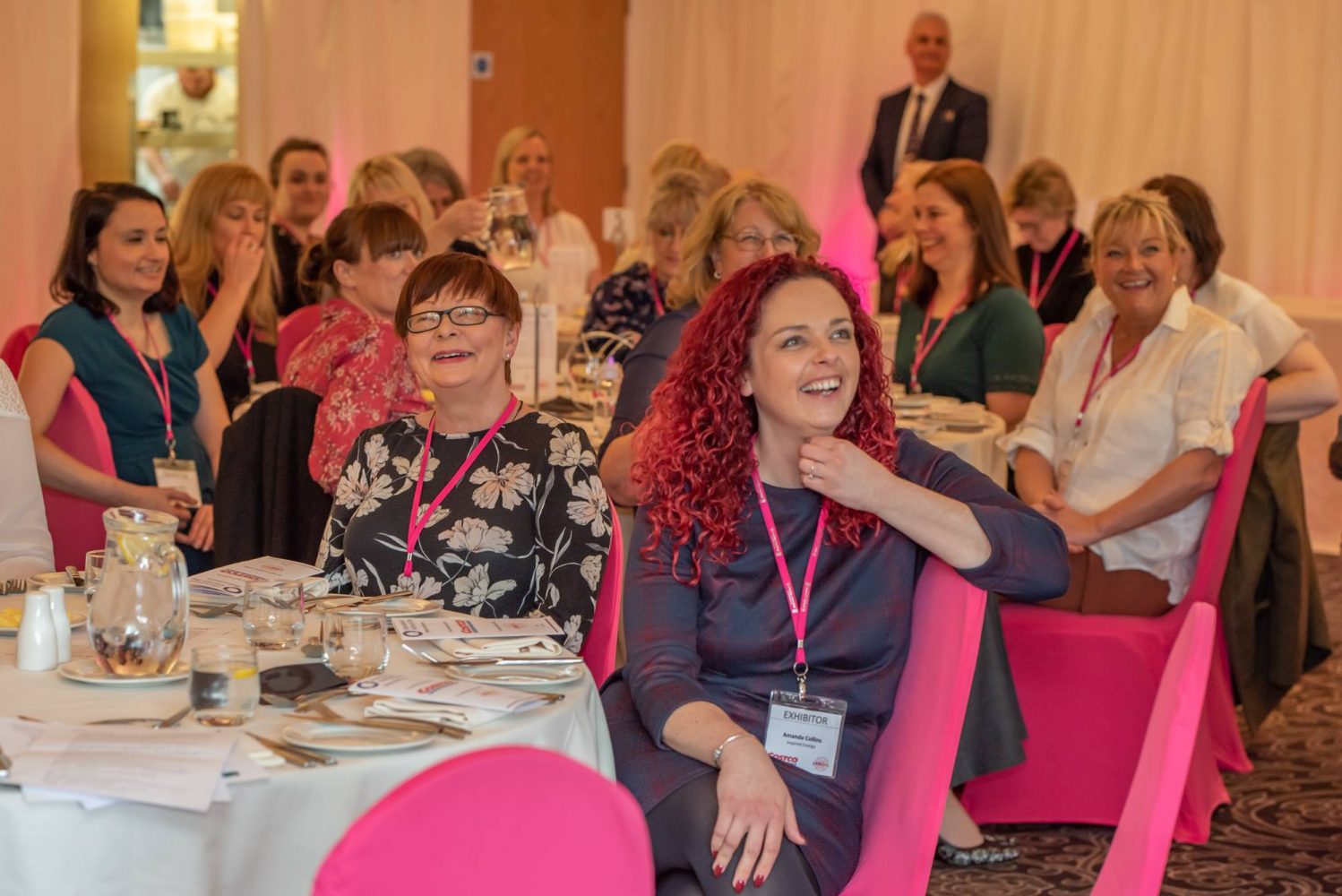 Large group of ladies sitting on pink covered chairs. Lady in the foreground has pink curly long hair and a purple dress on.